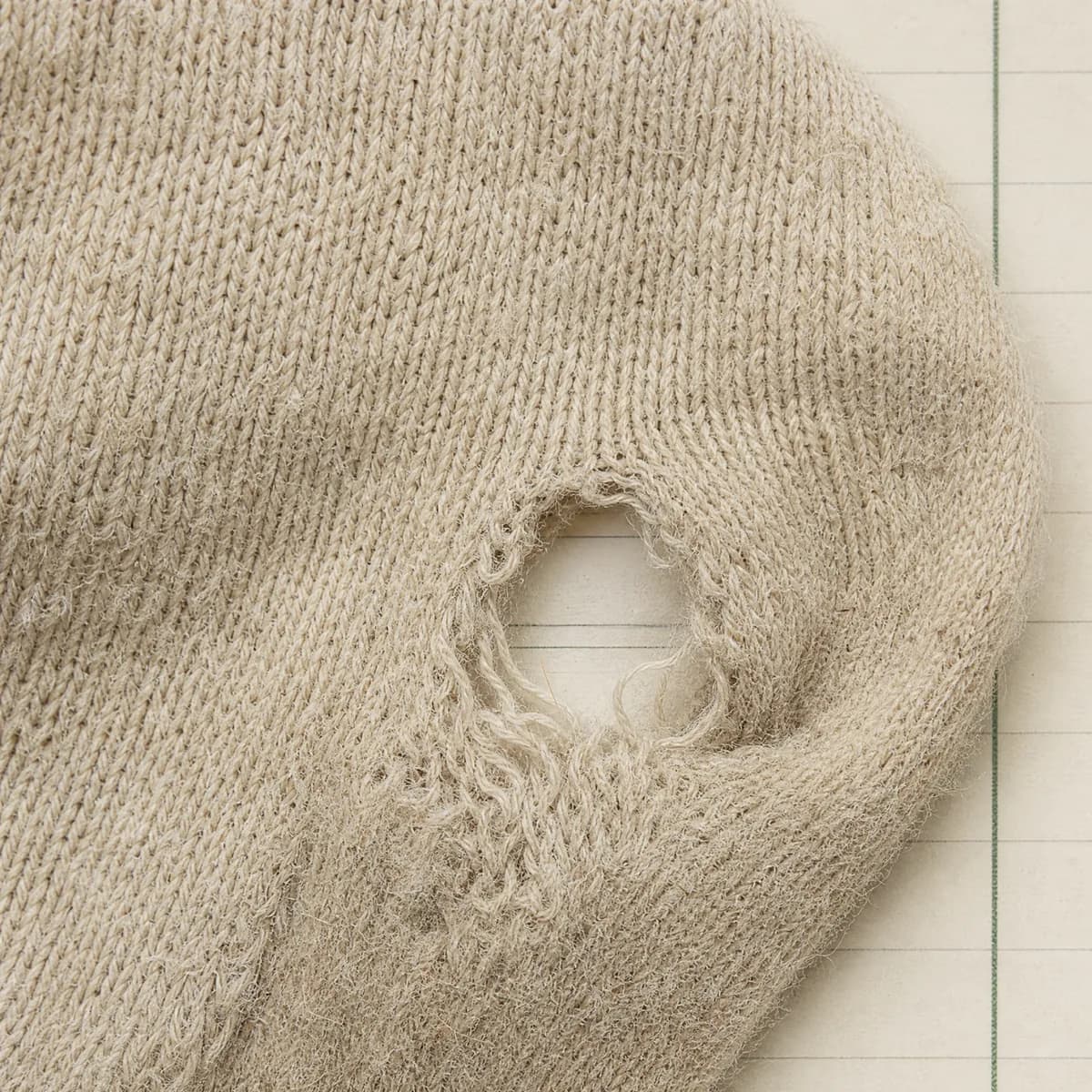 Extreme close-up of a small hole opening at the toe of a sock, the ruled paper visible through the gap.